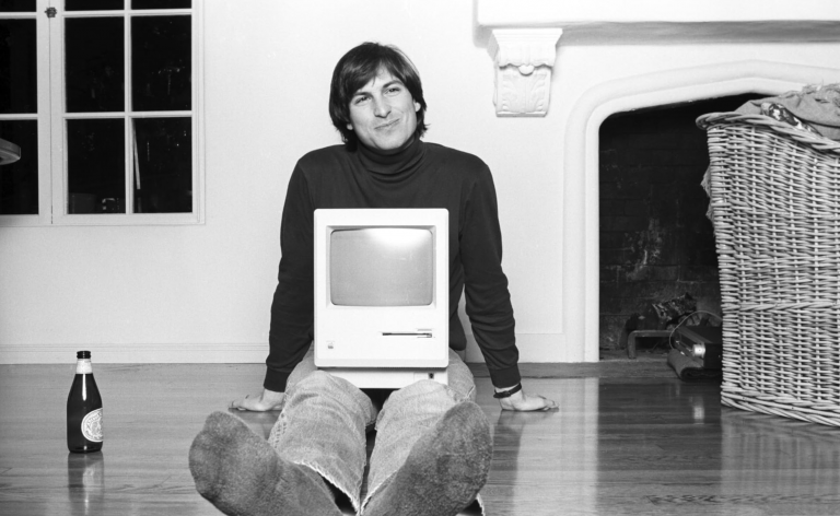 Black and white photo of Steve Jobs sitting cross-legged on the floor with the first Macintosh computer, smiling at the camera, in a room with a simple decor and a fireplace, evoking a moment of innovation and personal computing breakthrough.